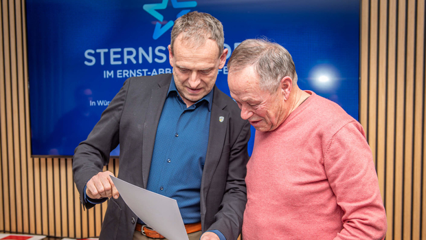 Zwei Männer betrachten ein Dokument vor einem Bildschirm mit dem Logo der Sternstunden