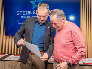 Zwei Männer betrachten ein Dokument vor einem Bildschirm mit dem Logo der Sternstunden