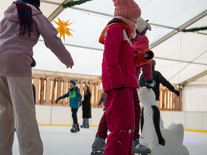 Kinder üben mit einer Eislaufhilfe auf der überdachten Eisbahn unter winterlicher Dekoration